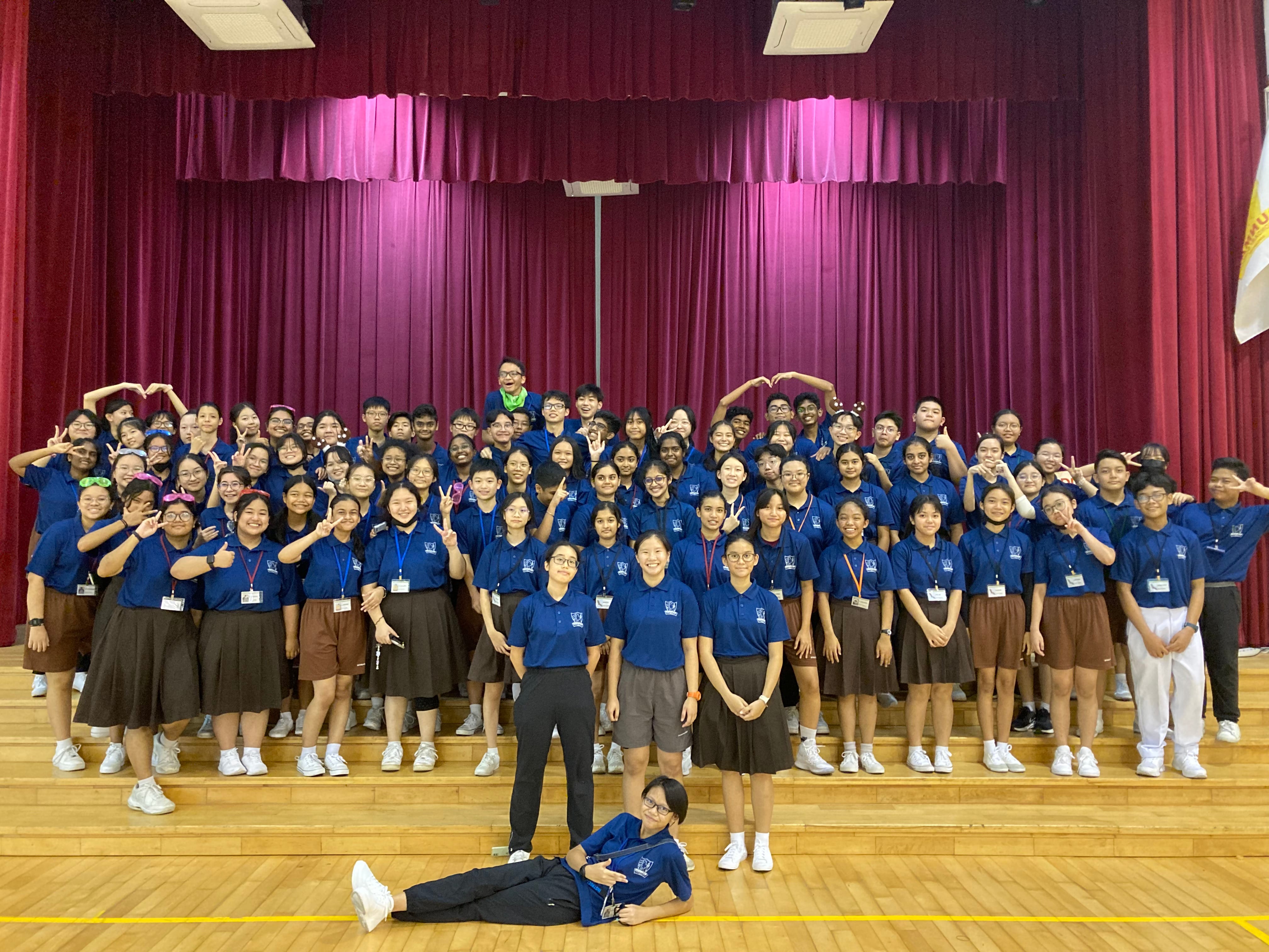 Large group of students in blue polo shirts posing with hand signs on a school stage with red curtains.